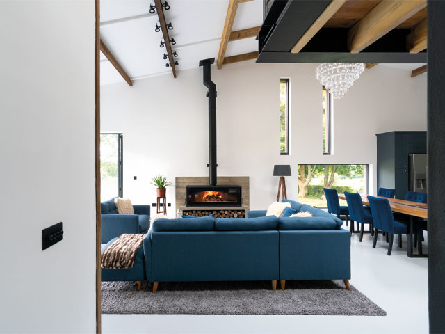 Grand Designs renovations. Double height ceiling large wood burning fire wooden dining table blue chairs large blue l-shaped sofa exposed wooden beams